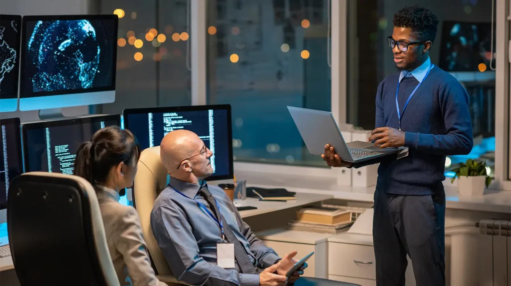 Man standing with laptop in front of two people sitting in front of computer monitors